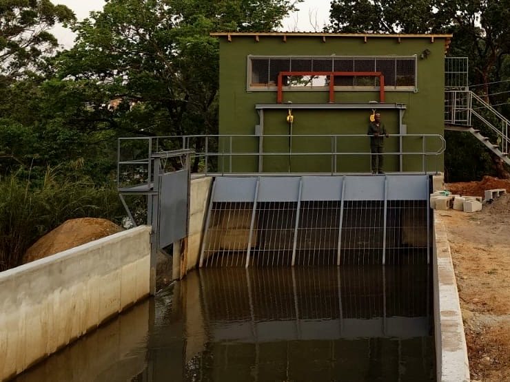 Intake structure of a run-of-river hydro scheme: steel trash racks across a concrete headrace, water held behind them, powerhouse above.