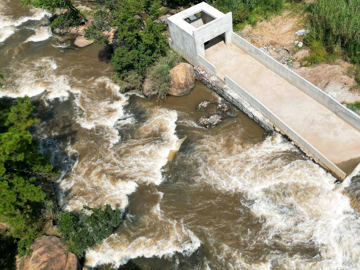 Aerial view of a river in full flow beside a concrete run-of-river intake and spillway, dense bush along both banks.