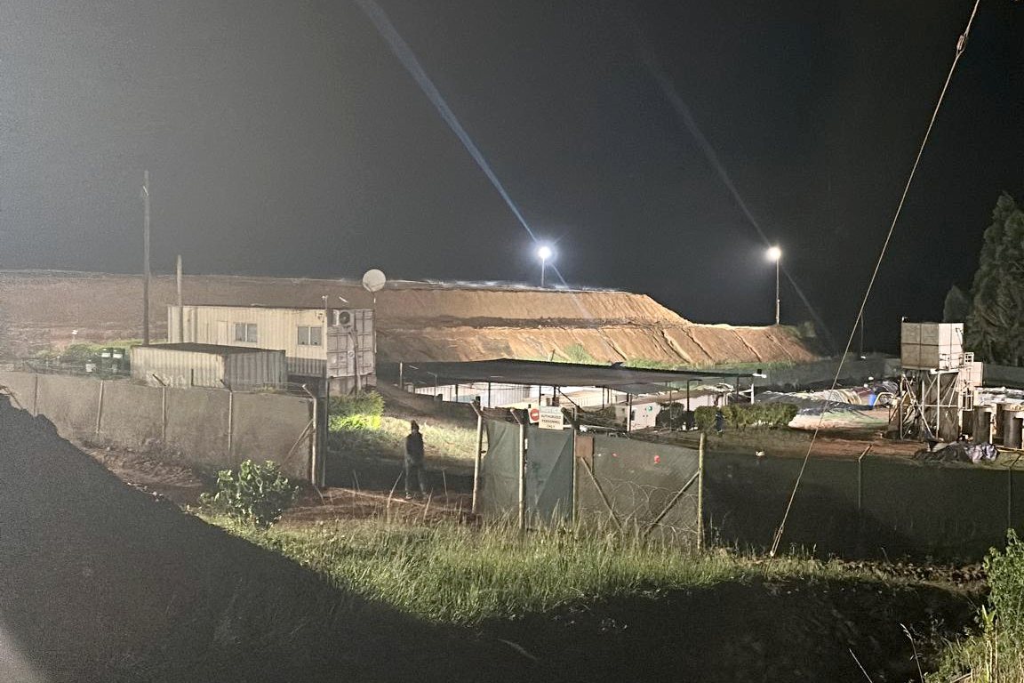 Mine compound at night: site offices and containers, a lit waste dump behind, plant and equipment to the right.